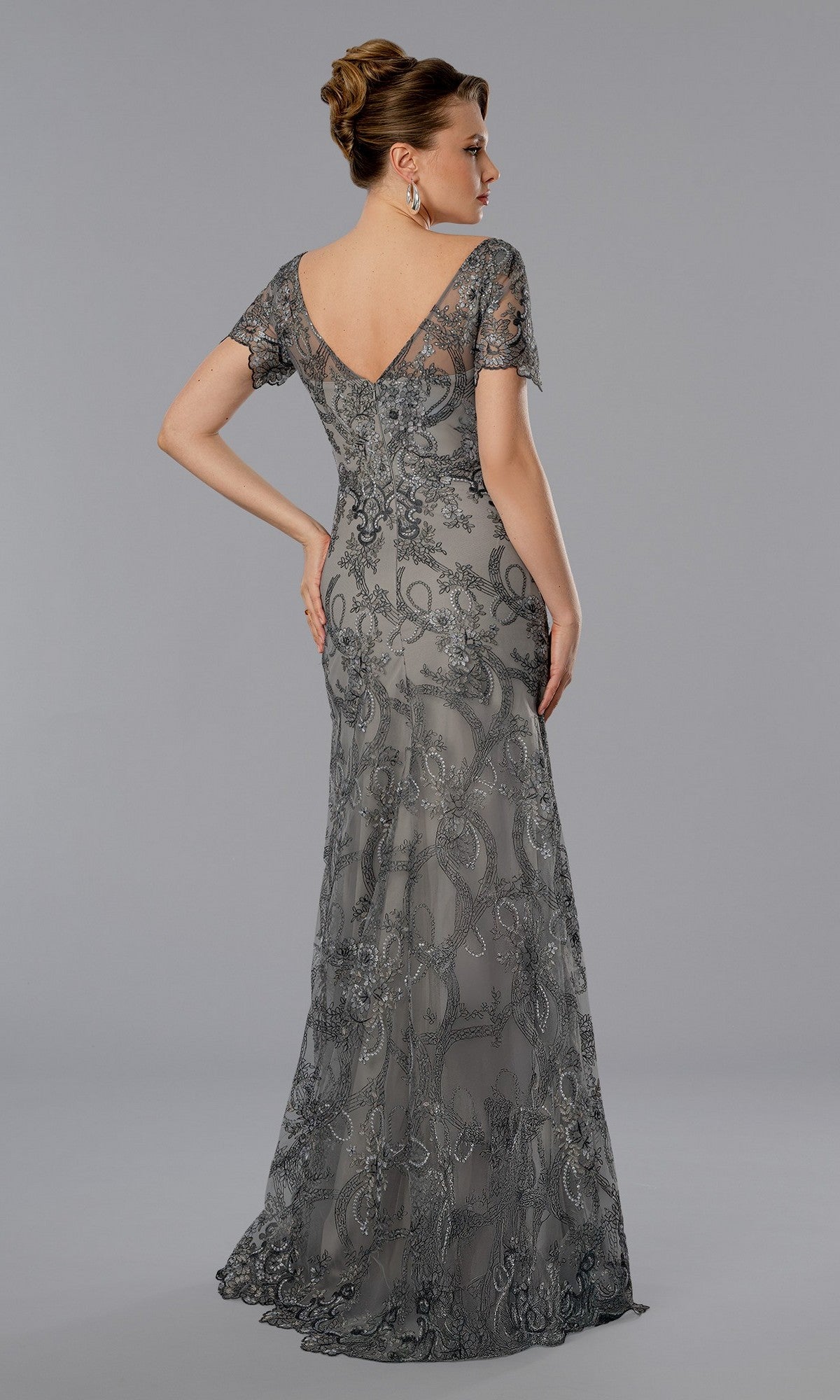 A woman models the Stella Couture 25369 long formal dress, featuring a silver-gray floral lace overlay, sheer short sleeves, and an elegant floor-length design, shown from the back against a plain gray background.